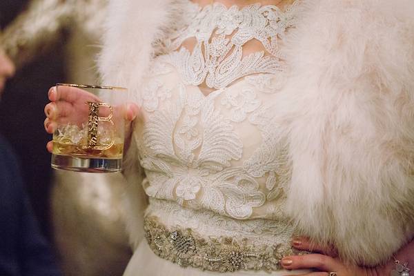 Close up of intricate lace Art Deco gown and a hand holding a golden plated cocktail glass