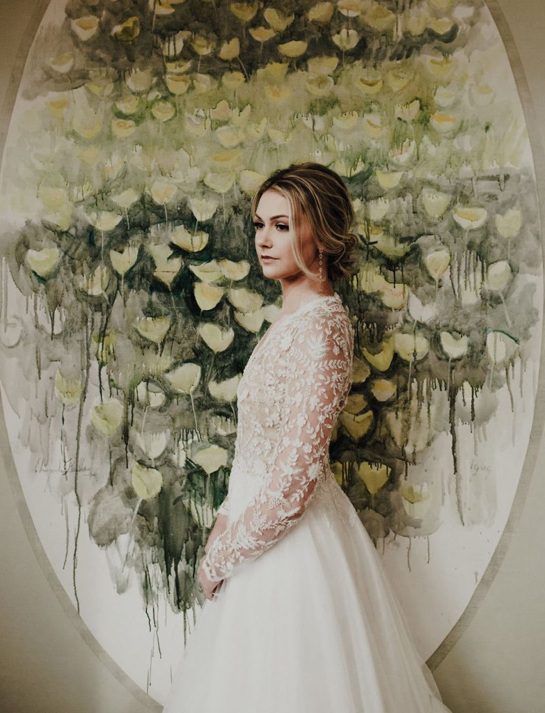 Woman standing in front of a green and gold painting wearing a white wedding gown with lace sleeves