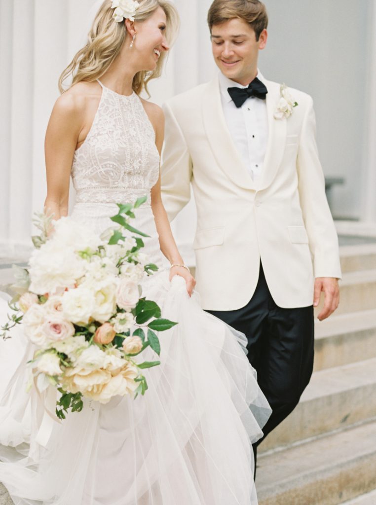 Bride and groom standing together. Bride is in a white halter gown and groom is in a cream tuxedo with black bow tie.