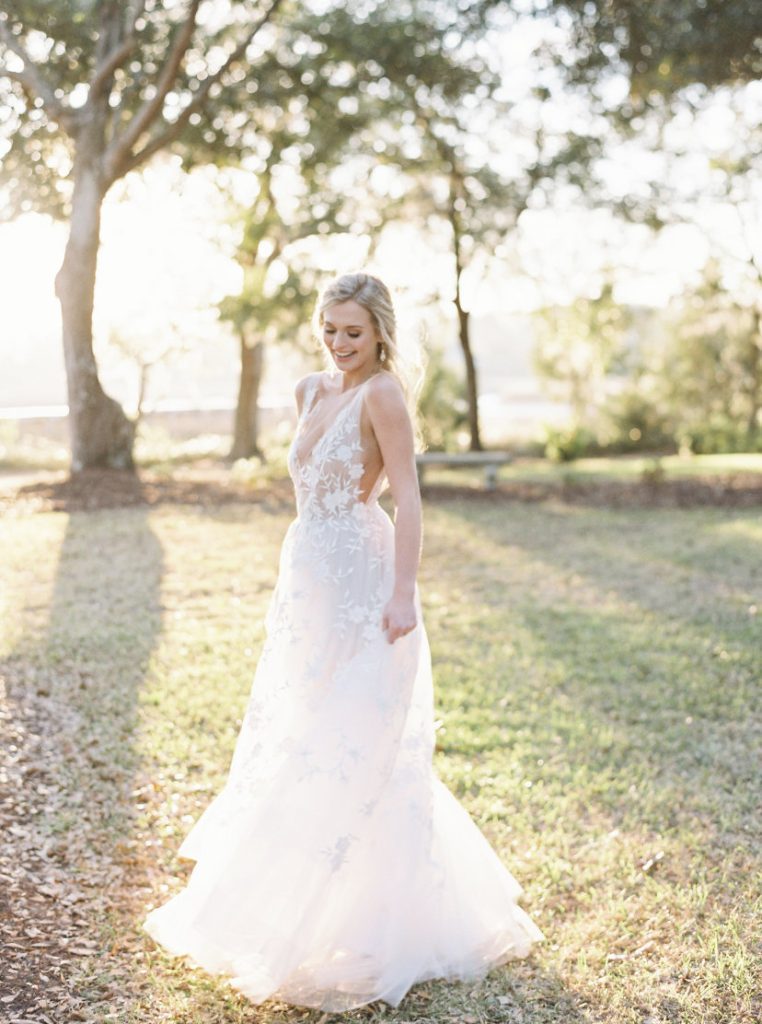 Bride standing in a small field surrounded by trees. Her dress has a fabric flowered top with a deep neckline.