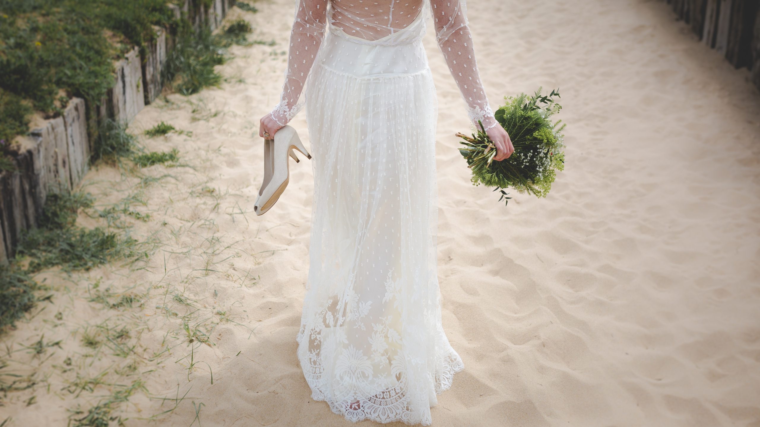the back of a woman walking on the beach wearing a wedding dress and holding her shoes in her hand