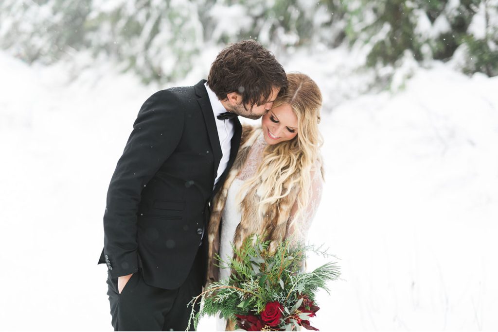 bride and groom in the snow. Bride wearing a lace white gown with a fur vest over it and carrying red roses