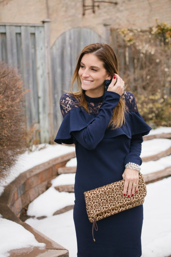 woman standing in the snow wearing a navy blue long sleeved dress with lace neckline and a leopard print clutch purse
