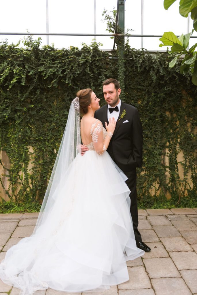 Bride and groom in a botanical garden. Groom is wearing a tux and a bow tie. Bride is wearing a tulle skirt and a lace long sleeved bodice.