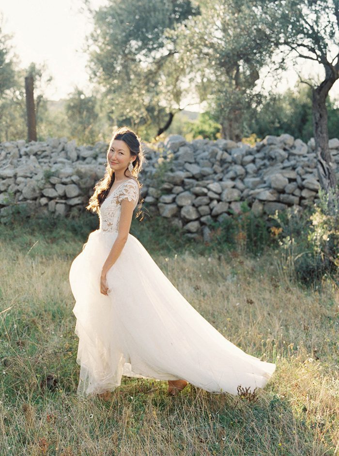 Bride standing in a field with a stone wall behind her. The dress has floral lace on the top and a tulle skirt