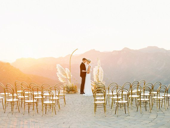 Bride and groom standing under a wedding arch with mountains in the background at sunset.