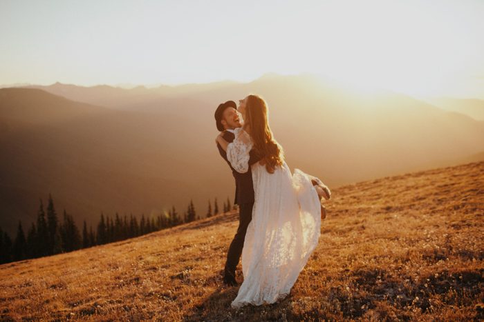 Groom lifting a bride up and swinging her around in a field surrounded by misty mountains at sunset